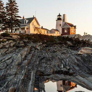 Pemaquid Point Lighthouse and keeper’s house in Bristol, Maine, glowing at sunrise and reflected in a tide pool on the rocky Atlantic shoreline.