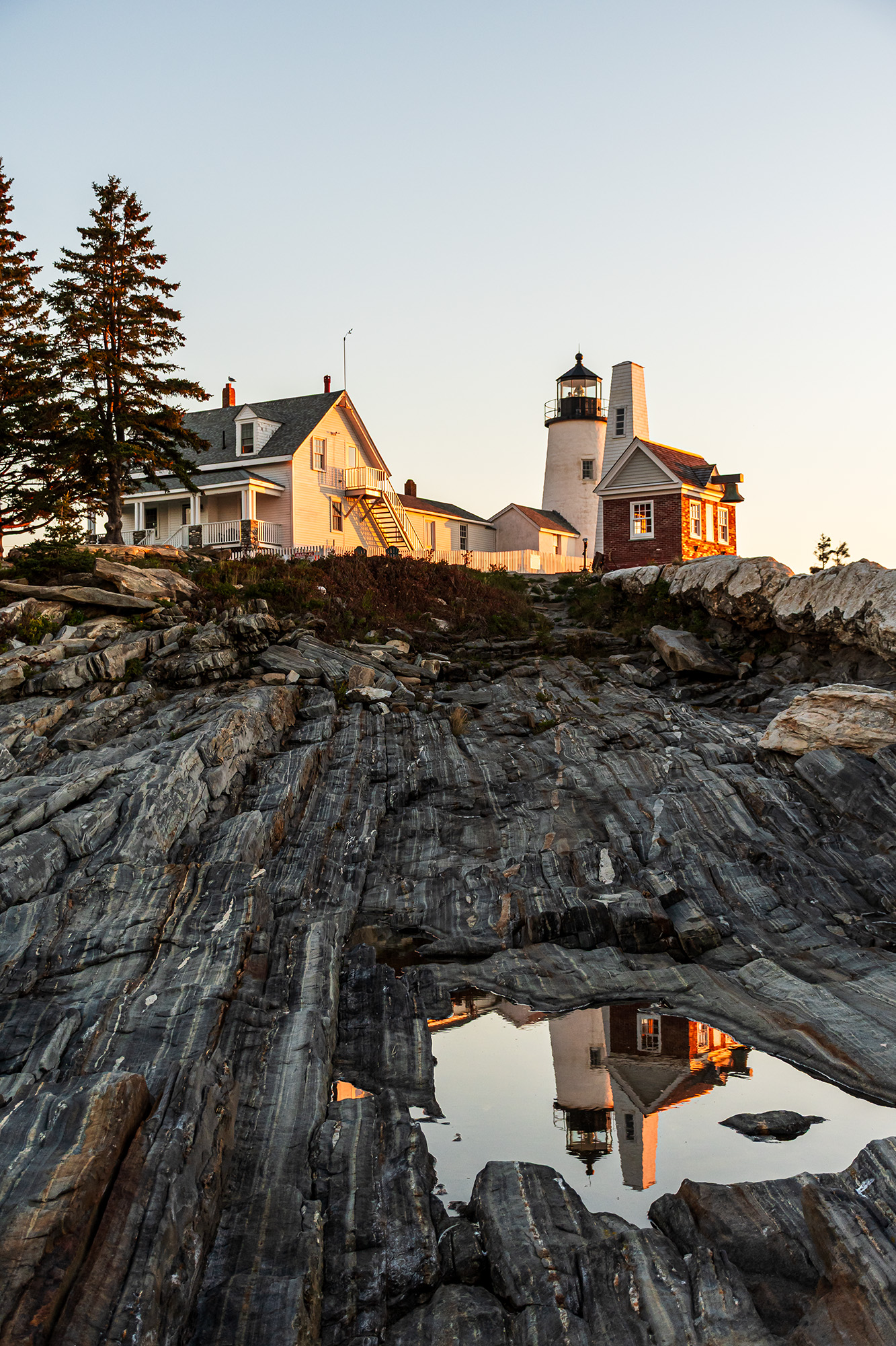 Pemaquid Point Lighthouse and keeper’s house in Bristol, Maine, glowing at sunrise and reflected in a tide pool on the rocky Atlantic shoreline.
