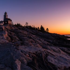 Pemaquid Point Lighthouse at sunrise, silhouetted against an orange and violet sky with granite ledges leading to the Atlantic Ocean in Bristol, Maine.