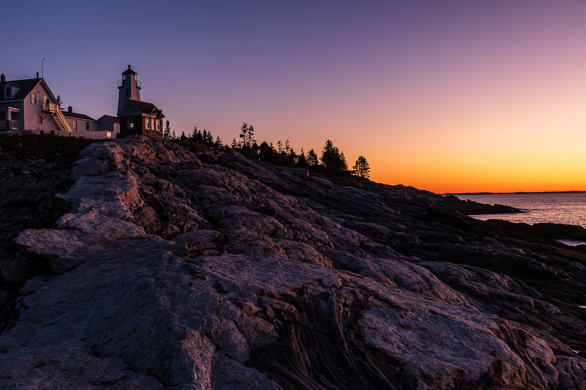 Pemaquid Point Lighthouse at sunrise, silhouetted against an orange and violet sky with granite ledges leading to the Atlantic Ocean in Bristol, Maine.