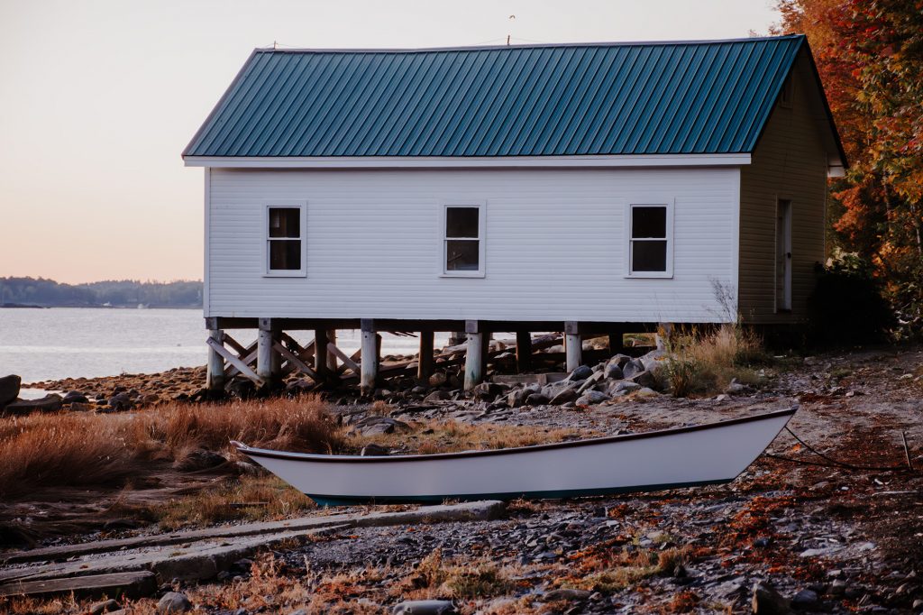 White boathouse on stilts with a small skiff resting on the shore at low tide in Northport, Maine.