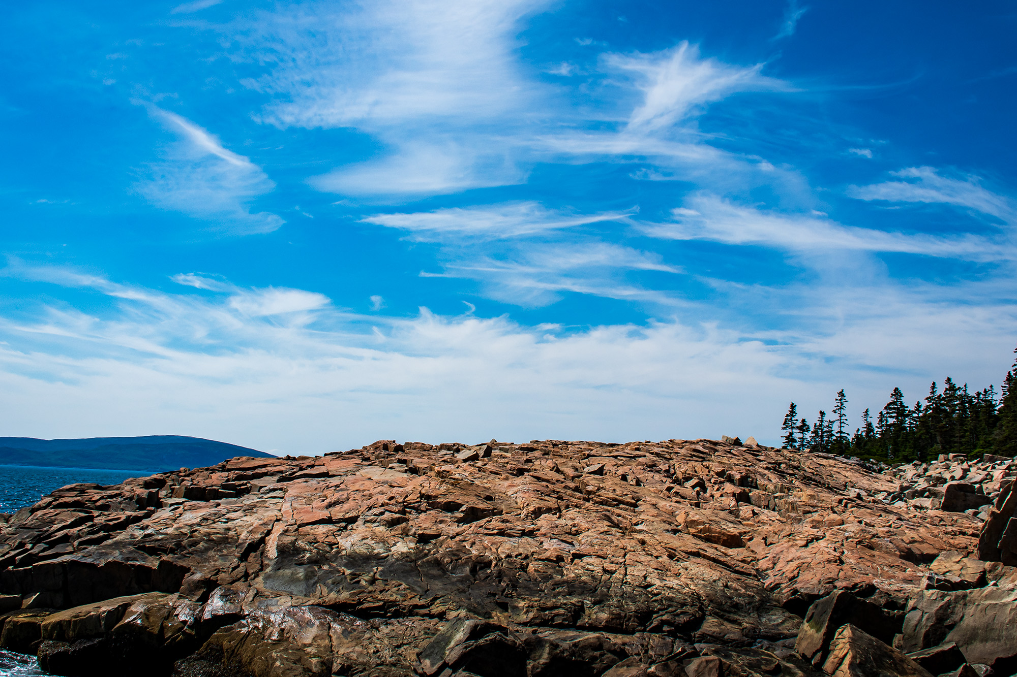 Granite ledges at Schoodic Point in Acadia National Park, Maine, beneath a bright blue summer sky with wispy clouds.