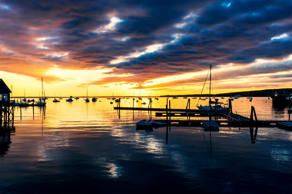 Sunrise over Rockland Harbor, Maine, with boats reflecting in calm golden water under dramatic morning clouds.