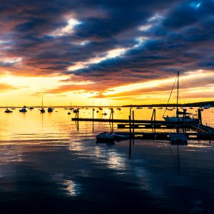 Sunrise over Rockland Harbor, Maine, with boats reflecting in calm golden water under dramatic morning clouds.