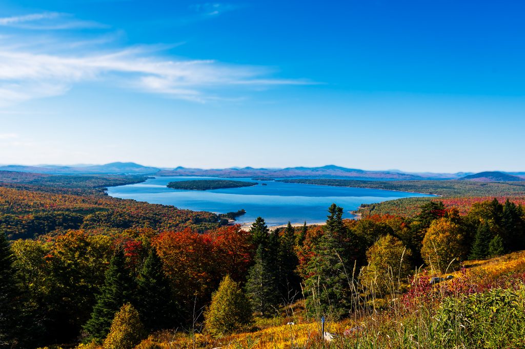 View from Height of Land in Western Maine, overlooking Mooselookmeguntic Lake surrounded by vibrant autumn foliage and distant mountain ranges under a clear blue sky.