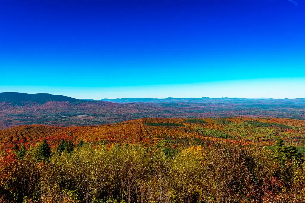 Expansive view from Quill Hill in Western Maine, showing mountains and valleys covered in peak autumn foliage beneath a vivid blue sky.
