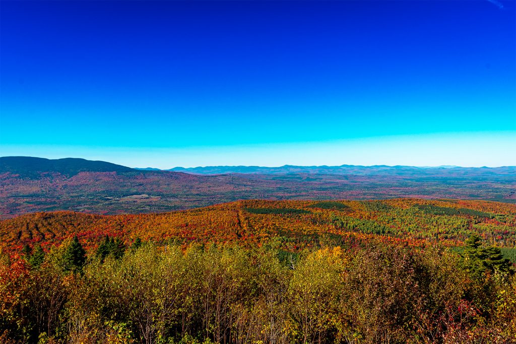Wide view of Western Maine mountains and forests covered in colorful fall foliage beneath a deep blue sky.