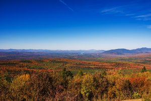 Expansive autumn landscape from Quill Hill in western Maine with colorful forests, distant lakes, and a bright blue sky.