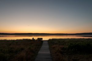 Wooden boardwalk leading to calm sunrise reflections over Sandy Point, Maine, with warm orange sky and still water.