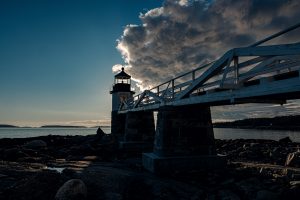 Marshall Point Lighthouse illuminated from behind by golden-hour light, dramatic clouds overhead, rocky shoreline in the foreground.