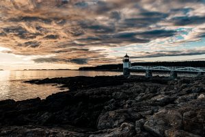 Marshall Point Lighthouse during late afternoon golden hour with dramatic clouds over rocky Maine shoreline.