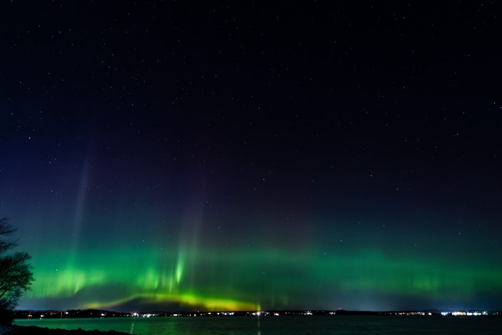 Northern lights glowing green over coastal waters and distant town lights in Bayside, Maine.