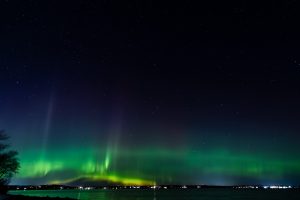 Northern lights glowing green over coastal waters and distant town lights in Bayside, Maine.