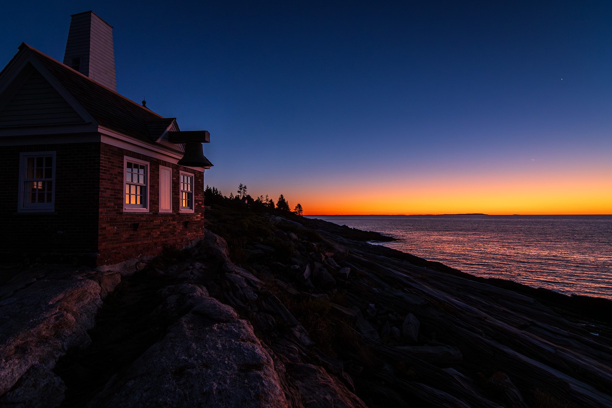Predawn light glows orange over the Atlantic Ocean beside Pemaquid Point Lighthouse in Bristol, Maine.